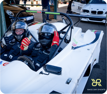 2 people sitting in a racing car, giving a thumbs up to the camera at a Race Day Event